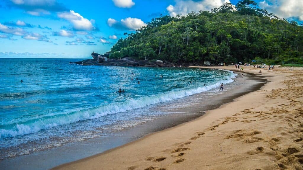 Ondas batendo na praia. Várias pessoas caminham na areia e se banham no mar, e ao fundo há uma colina repleta de vegetação.