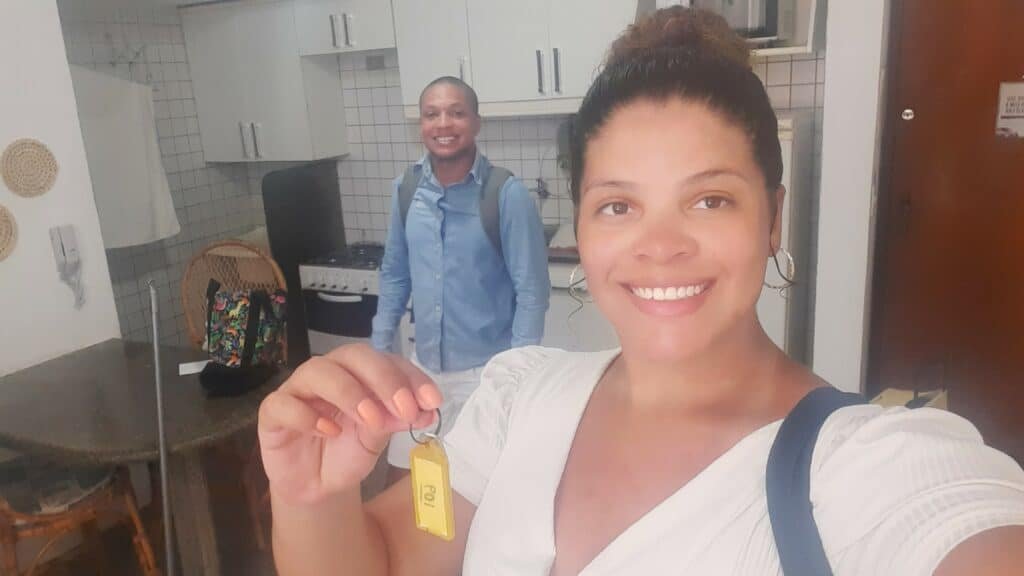 Selfie de uma mulher vestida de branco segurando uma chave com uma mão um homem atrás sorrindo com mochila nas costas. Eles estão dentro de um apartamento, com a pia da cozinha e mesa atrás. Representa Airbnb