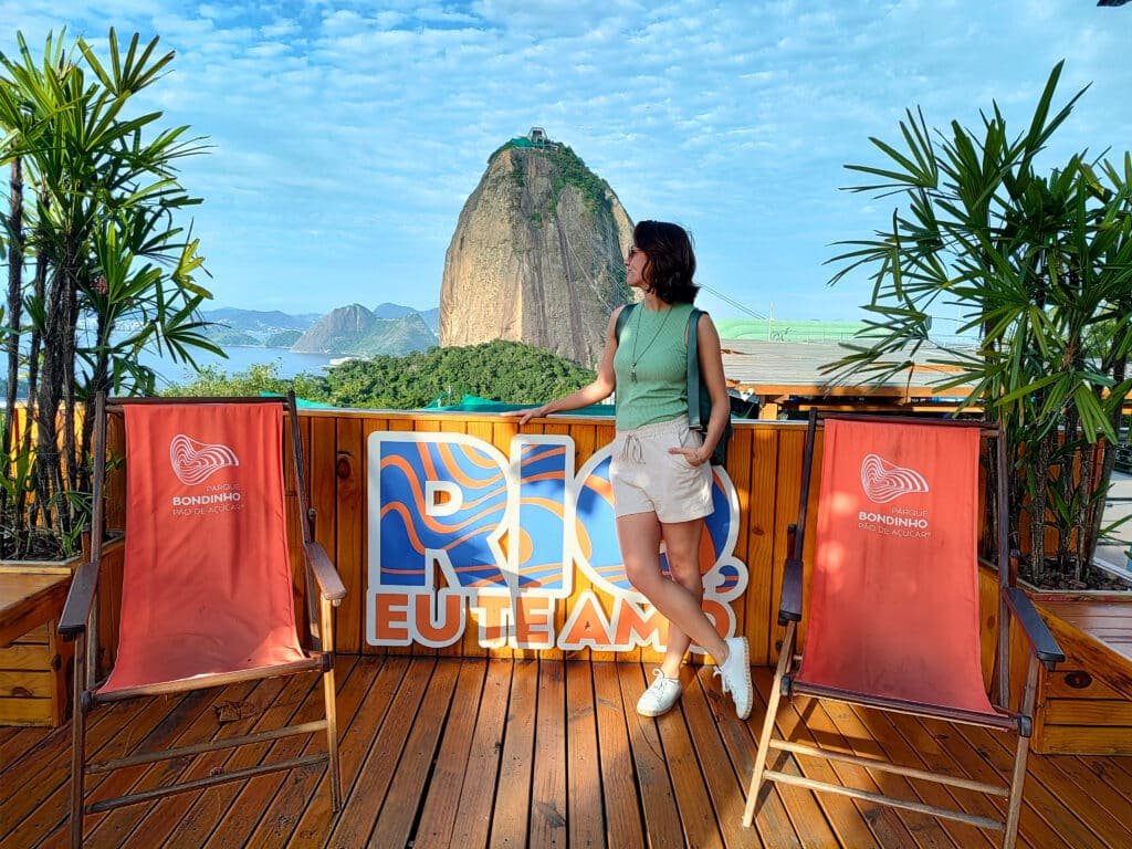 mulher posando para a câmera em frente ao corcovado no RIo de Janeiro. Há duas cadeiras de praia vermelhas de cada lado da moça, ela está olhando para a esquerda, permitindo aparecer somente o perfil do rosto