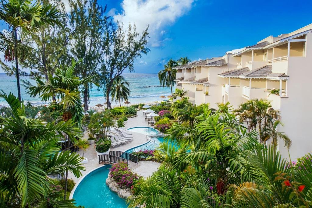 Parte externa do Bougainvillea Barbados que mostra várias árvores verdes e algumas plantas coloridas. Na parte de baixo da imagem tem uma piscina em formato de rio, do lado direito tem a construção do hotel e ao fundo o mar.