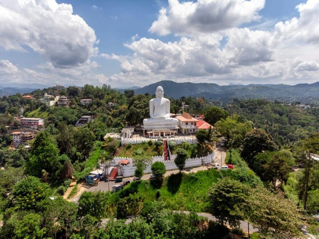 Uma grande estátua branca de Buda no topo de uma colina cercada por árvores e edifícios sob um céu parcialmente nublado, em Sri Maha Bodhi Viharaya. Representa eSIM Sri Lanka.