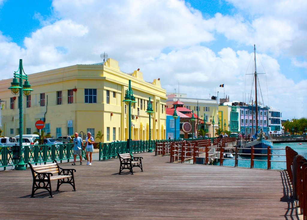Calçadão de madeira com bancos à beira da água, edifícios coloridos, pessoas caminhando e um veleiro atracado sob um céu azul. Imagem para ilustrar post sobre eSIM Barbados.