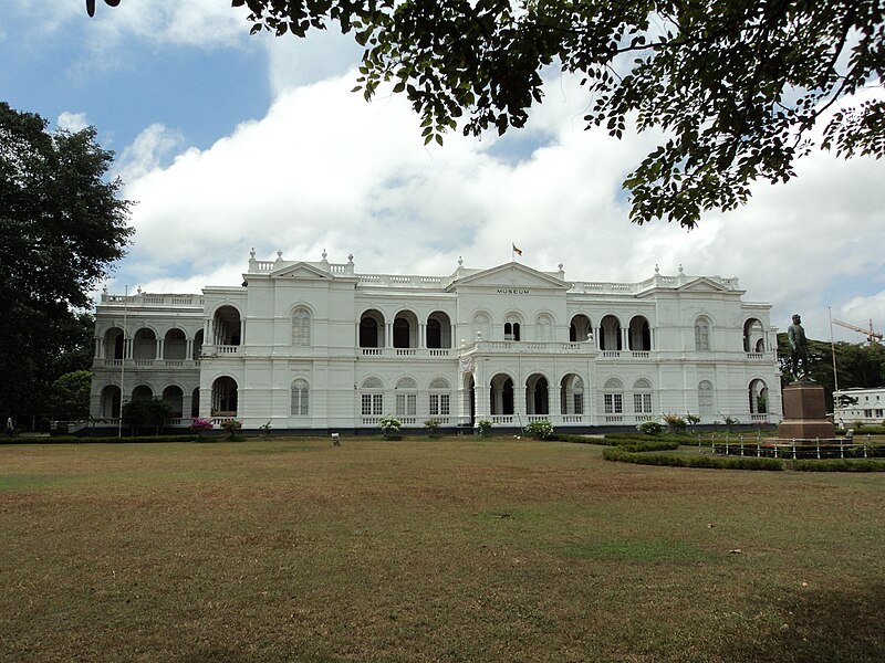 Grande edifício branco em estilo colonial, com janelas e colunas em arco, situado contra um céu parcialmente nublado, com um gramado na frente do Colombo National Museu.