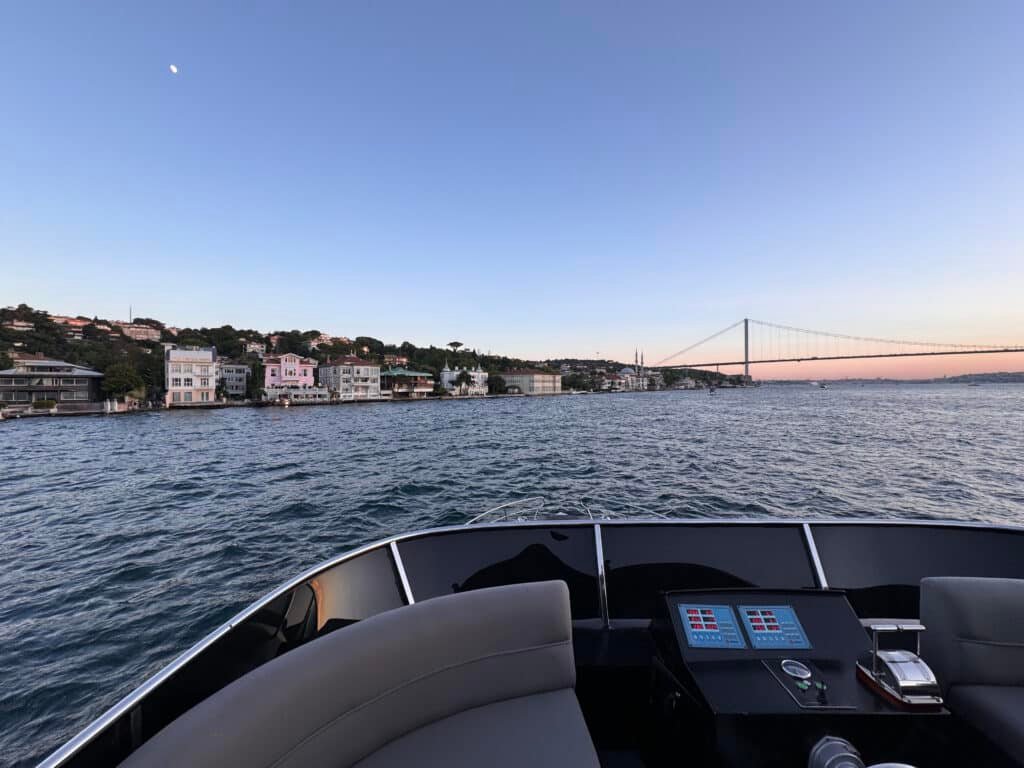 Vista do convés de um cruzeiro pelo Bósforo com edifícios ao longo da costa e uma ponte visível à distância sob um céu limpo.