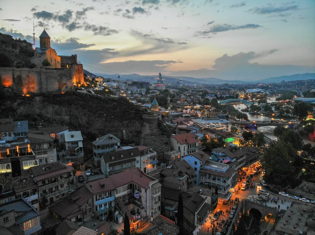 Uma vista áerea de Tiblissi, a capital da Georgia, com casas em estilo antigo que vão escalando uma montanha que no topo tem uma igreja, há duas pontes cortando a cidade e muitos carros pelas ruas