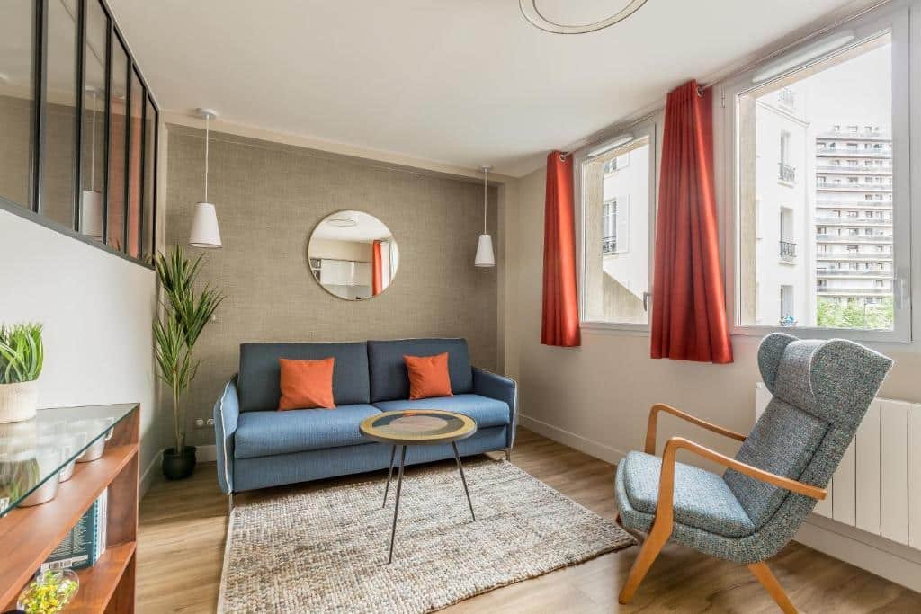 Sala de estar com uma poltrona e um sofá de dois lugares . Há uma janela à direita de vidro com vista dos prédios, e uma cortina laranja encolhida, além de uma mesinha de centro e um móvel com vaso de flor, llivros e canecas.