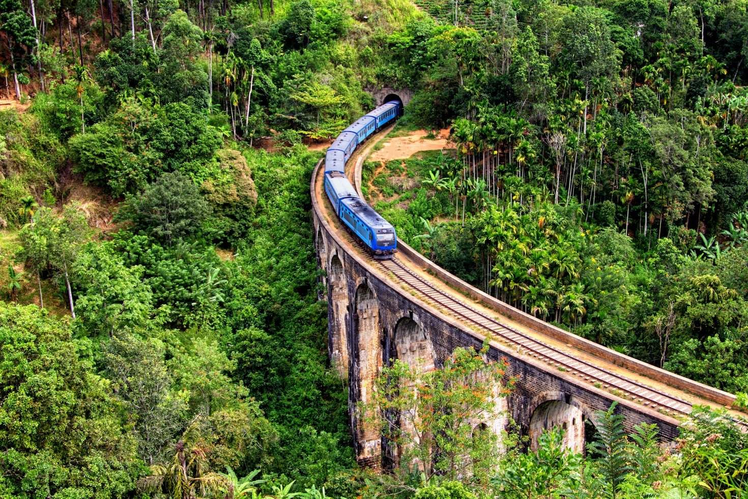 Um trem azul em Ella passa por uma ponte de pedra curva cercada por densa vegetação verde e árvores. Representa o que fazer no Sri Lanka.