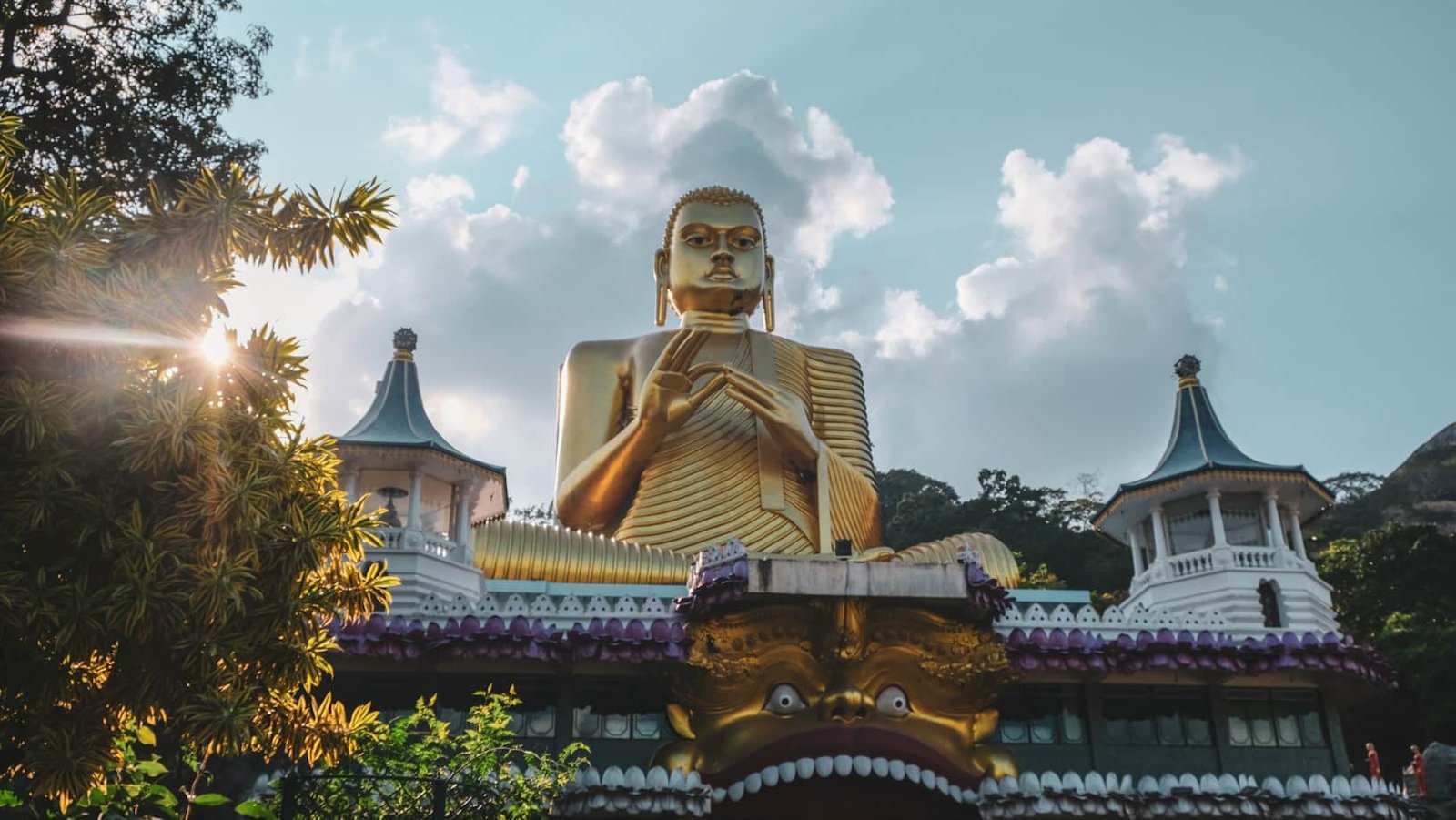 Uma grande estátua dourada de Buda no Golden Temple fica no topo de um edifício com fachada em formato de cabeça de leão, cercado por árvores e duas estruturas semelhantes a pagodes. Representa o que fazer no Sri Lanka.