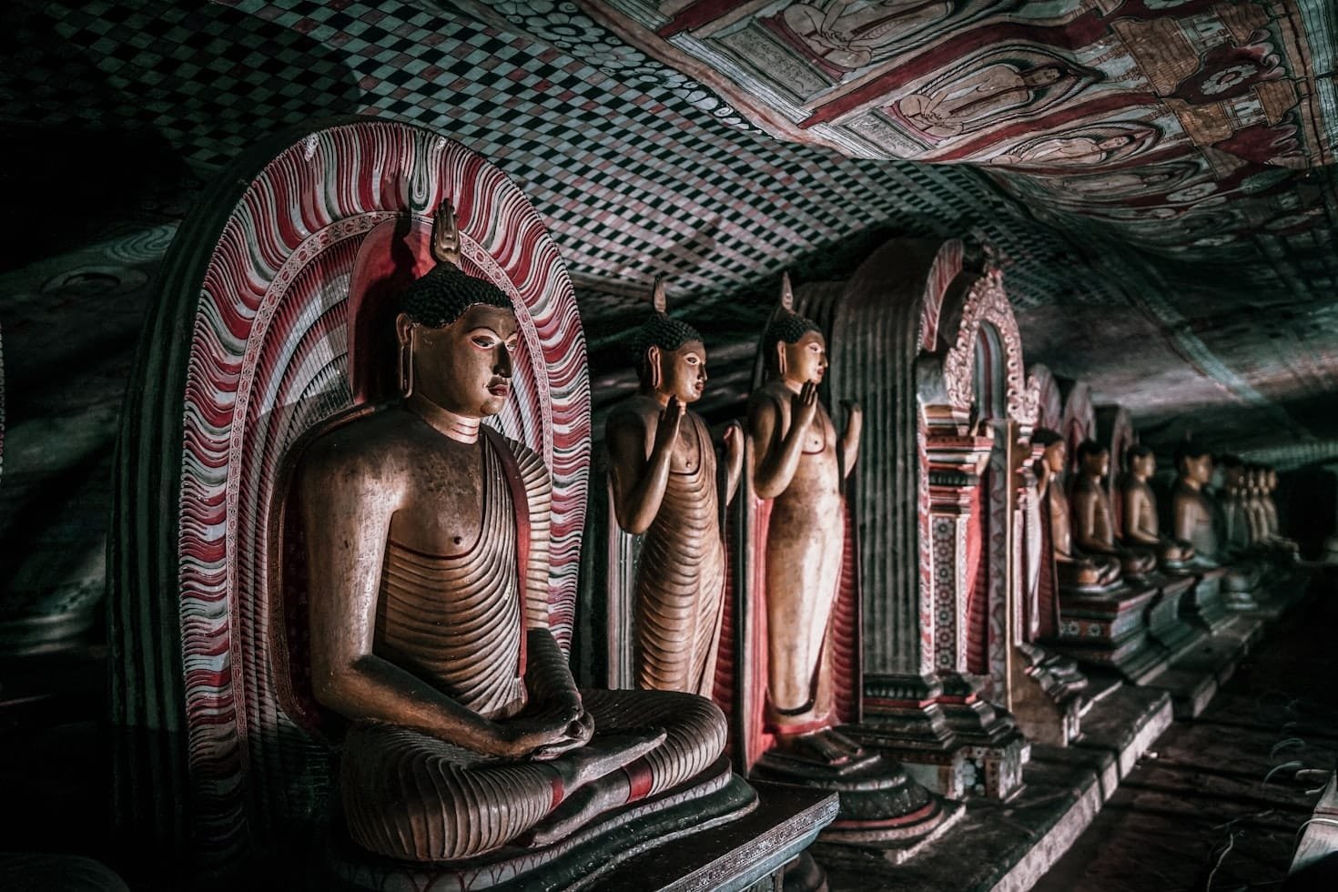 Uma fileira de estátuas de Buda sentadas e em pé em uma caverna mal iluminada com tetos e paredes pintados de forma complexa, em Dambulla.