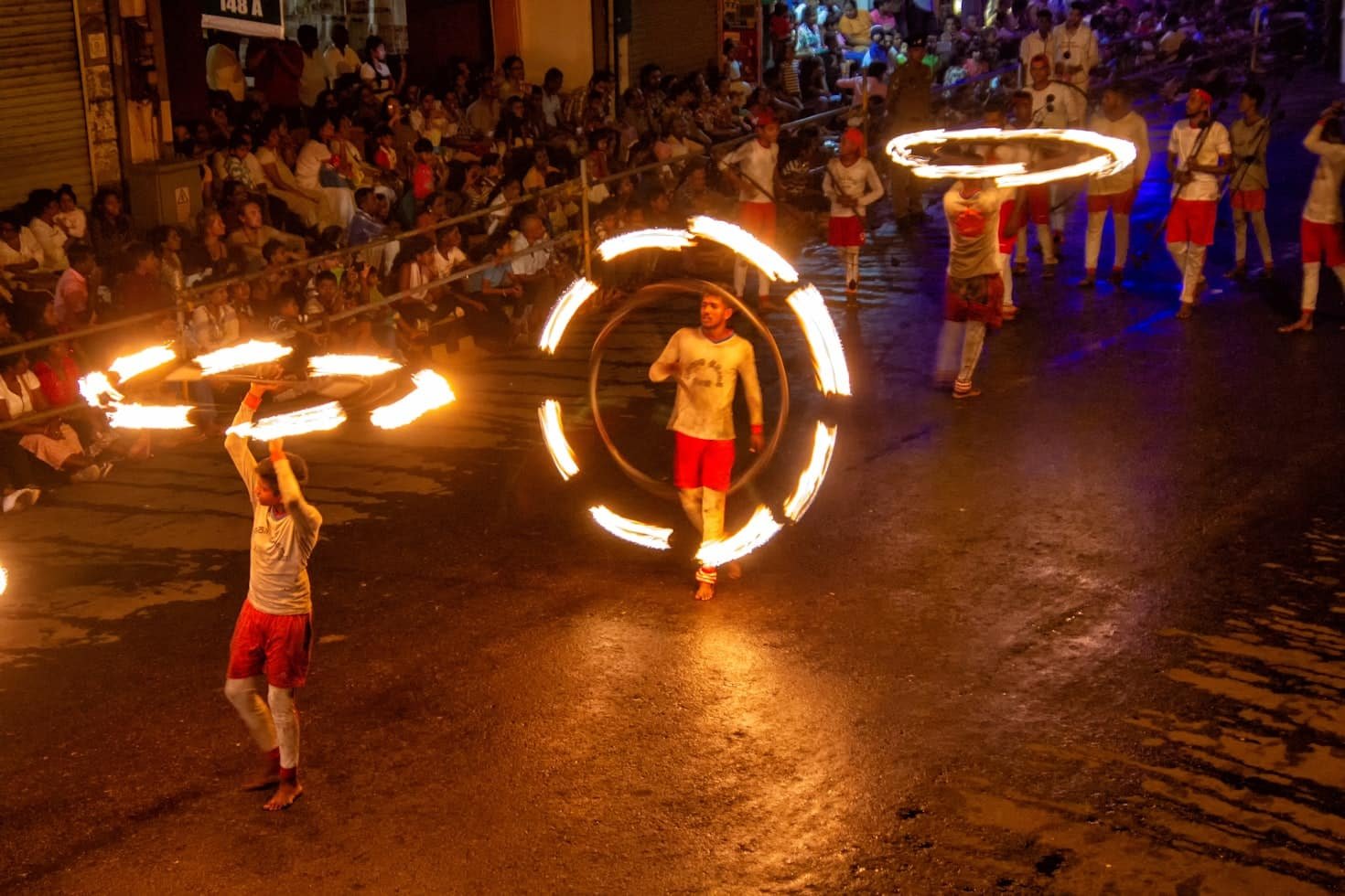 Artistas vestindo shorts vermelhos e camisas brancas em Kandy giram arcos em chamas em um desfile noturno, com uma grande multidão assistindo da calçada. Representa o que fazer no Sri Lanka.