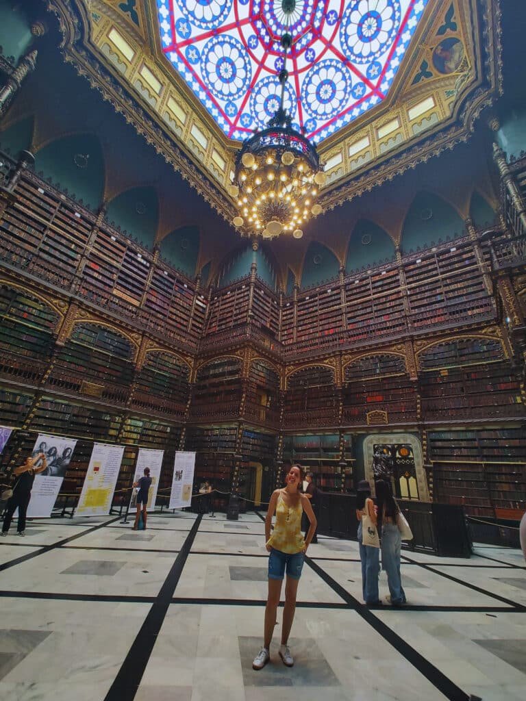 Interior de uma biblioteca magnífica com uma arquitetura detalhada e uma cúpula colorida de vitral no teto. As estantes altas e cheias de livros antigos cobrem as paredes, criando um ambiente impressionante e rico em história. No centro do salão, várias pessoas exploram o espaço, com uma jovem em destaque, vestindo uma blusa amarela e shorts jeans, olhando ao redor maravilhada. O chão de mármore branco e preto completa a elegância do local.