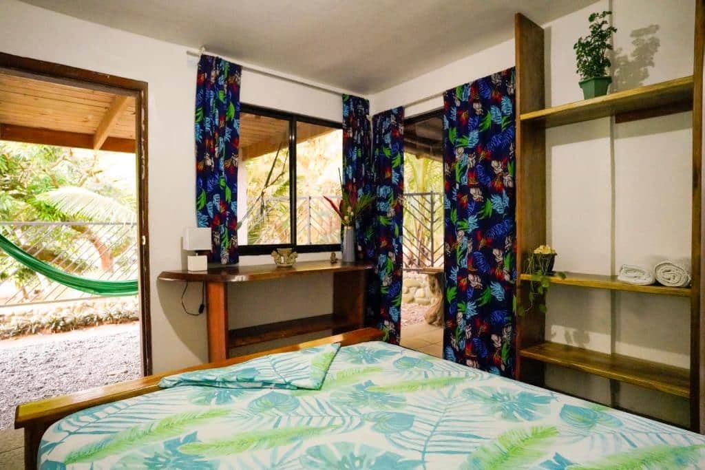 Um quarto com uma colcha com tema tropical, cortinas vibrantes, uma escrivaninha de madeira, uma varanda com rede e uma estante no Hotel Swell Pavones.