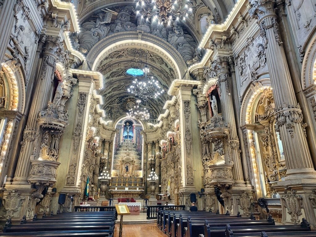 Um interior de igreja magnífico, com uma arquitetura barroca rica em detalhes. Arcos e colunas ornamentadas ladeiam o caminho até o altar, que é decorado com esculturas e entalhes intrincados. Lustres pendem do teto, iluminando o espaço com uma luz suave. O teto abobadado é adornado com relevos elaborados e pinturas, criando uma atmosfera de grandeza e reverência. Bancos de madeira escura estão dispostos em fileiras, conduzindo o olhar dos visitantes para o impressionante altar ao fundo