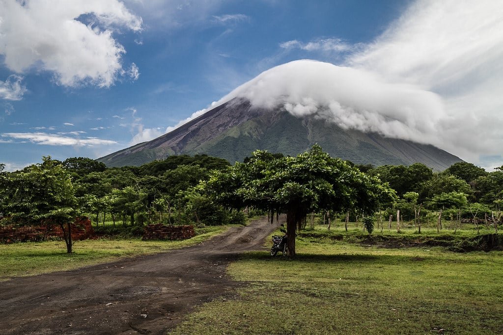 Uma estrada de terra leva a uma árvore em frente a um grande vulcão em Isla Ometepe com nuvens cobrindo seu pico sob um céu azul. Imagem para ilustrar post sobre eSIM Nicarágua.