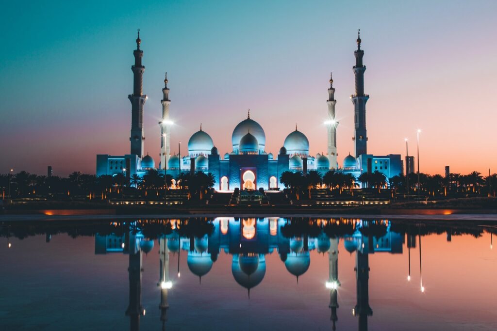 Mesquita branca iluminada por luzes azuis ao pôr do sol para ilustrar o post sobre chip de celular para Abu Dhabi. Um lago em frente à construção reflete a imagem da mesquita. - Foto: David Rodrigo via Unsplash