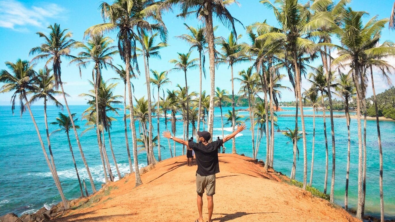 Pessoa em pé em um caminho de areia ladeado por altas palmeiras, de frente para o oceano, com os braços estendidos sob um céu azul claro, em Mirissa.