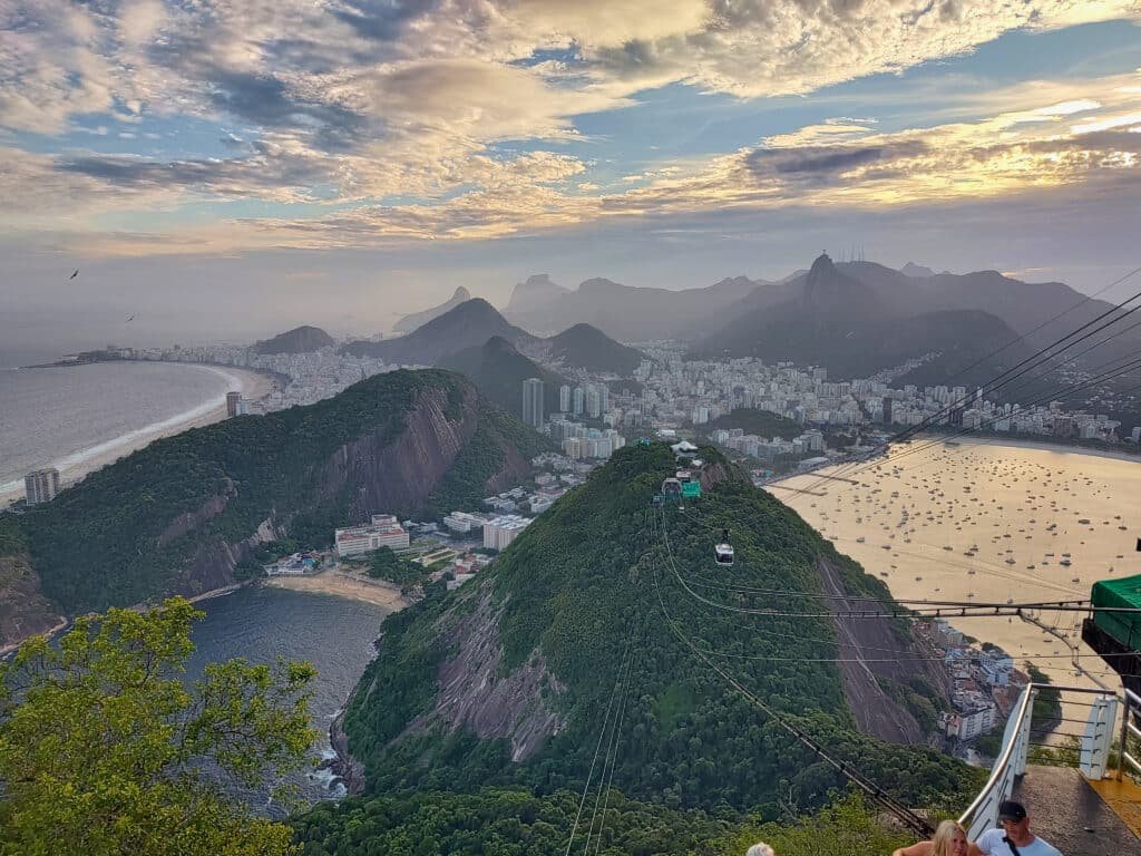 vista panorâmica do parque corcovado no alto do bondinho, no Rio de Janeiro. É possível enxergar o mar, os prédios, o contorno das montanhas e o pôr do sol ao fundo.