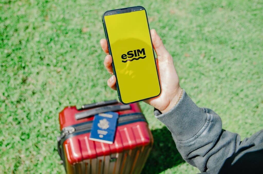 Uma pessoa segura um smartphone com a inscrição "eSIM" em uma tela amarela, acima de uma mala vermelha e um passaporte, na grama verde. Representa eSIM Bangkok.