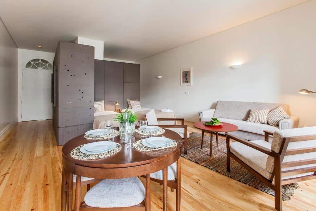 Sala de estar do bnapartments Loftpuzzle com mesa redonda de madeira a frente, do lado direito da imagem a sala de estar com duas poltronas e um sofá e ao fundo duas camas de solteiro.