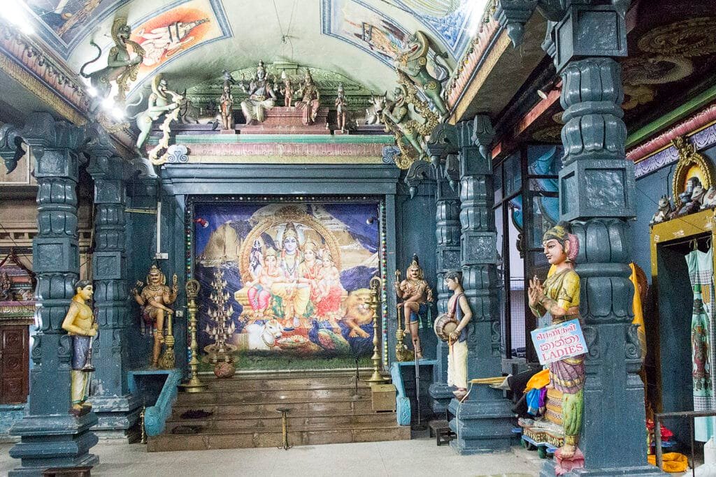 Interior de um templo hindu do Templo Sri Kailawasanathan Swami Devasthanam Kovil com pilares ornamentados, entalhes e um mural colorido representando divindades. Estátuas enfileiram-se nas laterais.
