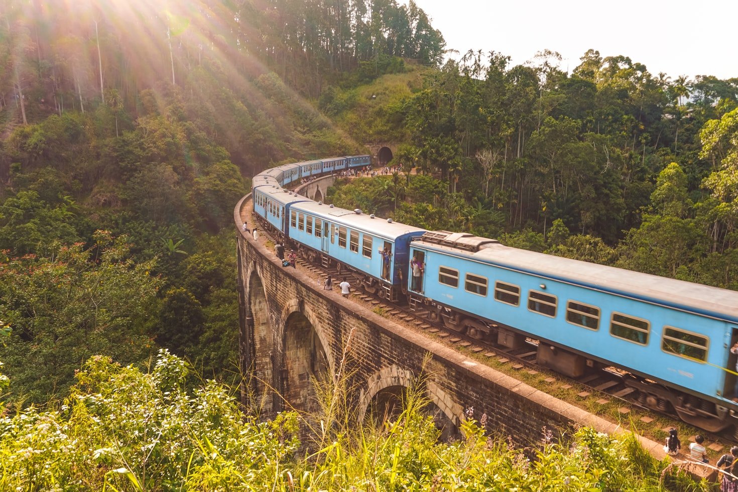 Um trem azul viaja em uma ponte de pedra em Ella curva através de uma floresta verdejante com a luz do sol entrando pelas árvores.