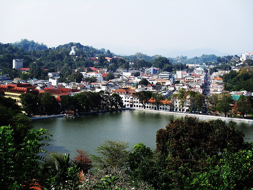 Vista aérea de uma cidade de Kandy com um grande lago em primeiro plano, cercada por edifícios, árvores e colinas sob um céu limpo. Representa o que fazer no Sri Lanka.